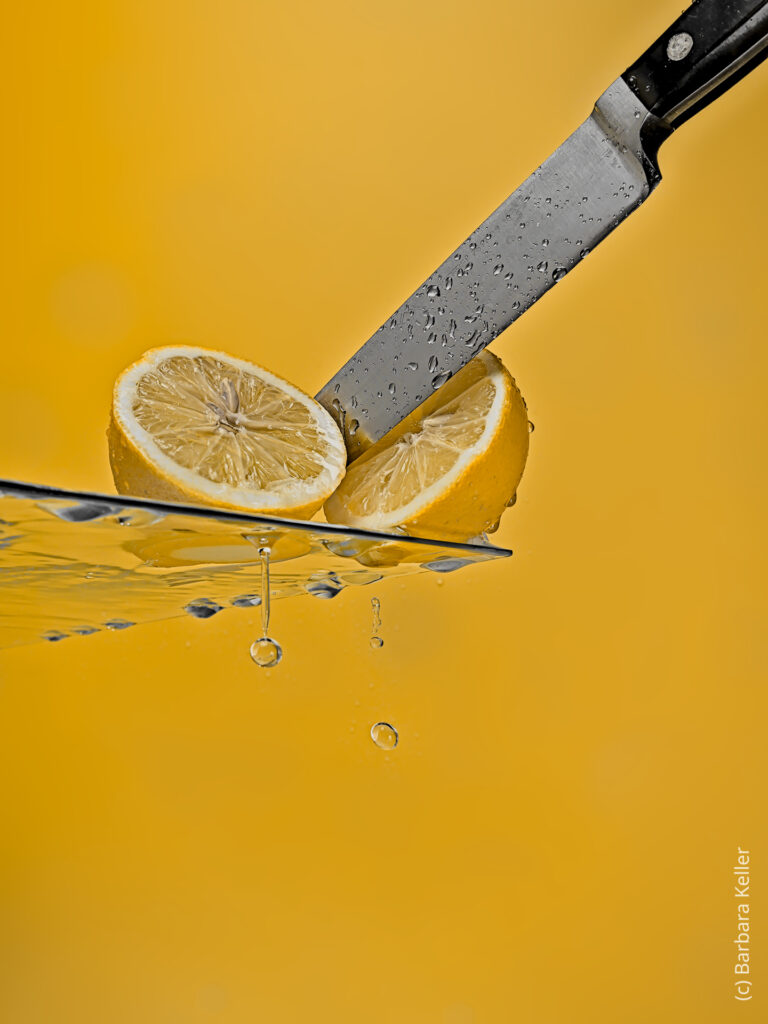 Produktfoto einer Zitrone die zerschnitten wird. Wasser tropft von der Glasplatte auf der die Zitrone liegt